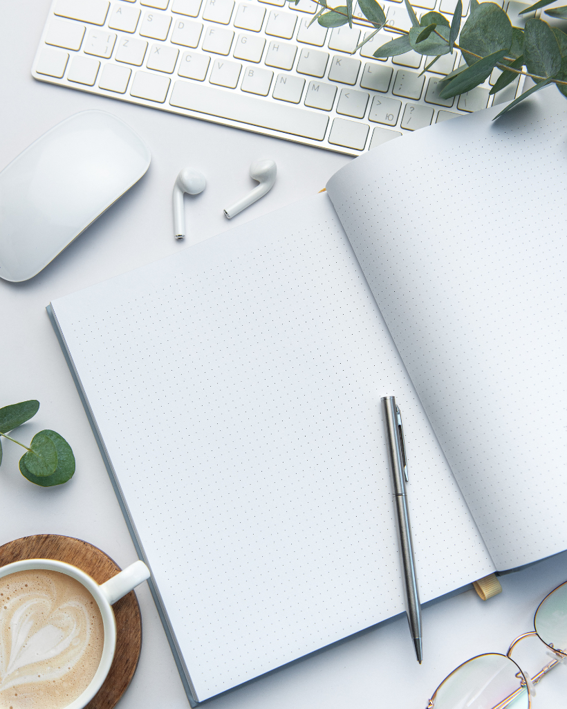 Open dotted notebook with a pen, wireless earbuds, keyboard, mouse, coffee cup, glasses, and green leaves on a white NY or FL-inspired desk.