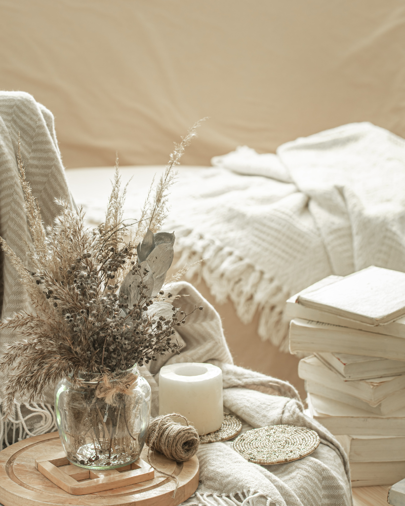 A glass vase with dried flowers, a candle, and stacked white books are styled on a table—evoking cozy NY and FL vibes with neutral blankets in the background.